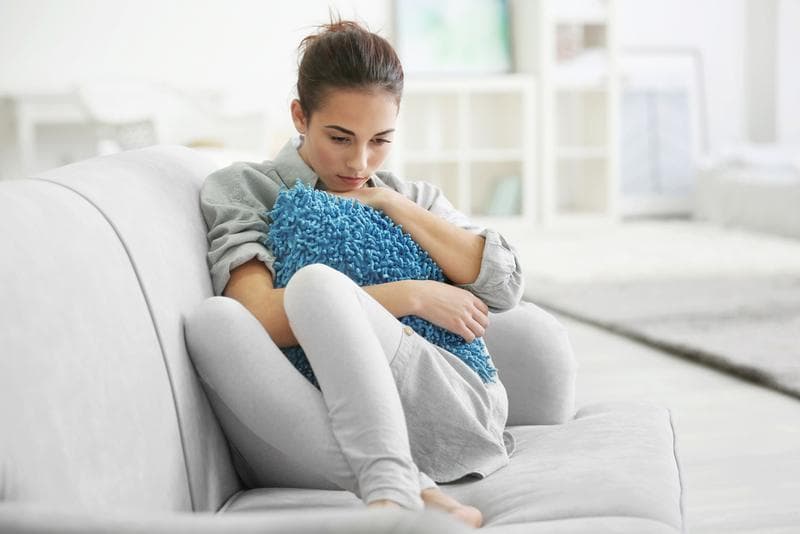 Woman sitting on a couch, hugging a pillow, appearing sad and withdrawn. 