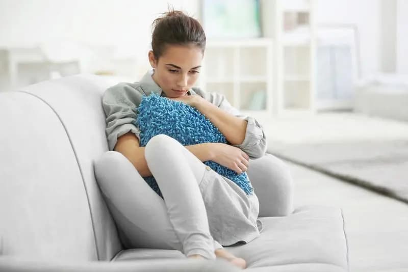 Woman sitting on a couch, hugging a pillow, appearing sad and withdrawn.